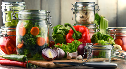Jars with marinated food and raw vegetables on cutting board