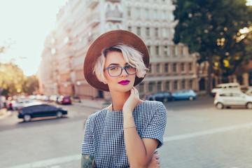 Portrait of pretty blonde girl with short hair posing to the camera on the steet in city. She wears gray checkered  dress, glasses, hat and has purple lips. - Powered by Adobe