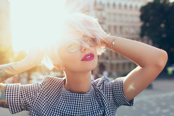 Closeup portrait of  blonde girl with short hair posing on the steet on sunset background. She wears gray checkered dress, glasses, has purple lips. Full of sunlight.