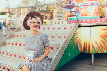 Cute blonde girl with short haircut is sitting on stairs of carousel. She wears checkered dress,  glasses, hat and has purple lips. She holds popcorn and smiles.