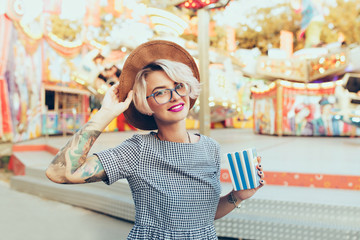 Obraz na płótnie Canvas Horizontal portrait of blonde girl with short haircut posing in amusement park. She wears checkered dress, glasses, hat and has purple lips. She holds popcorn and smiles.