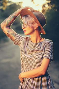 Vertical portrait of blonde girl with short haircut posing to  in forest on sunset background. She wears hat, sunglasses, gray checkered dress and has tattoo on hand.