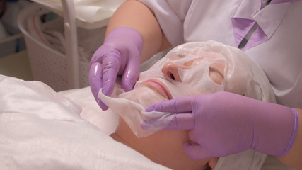 The procedure of skin rejuvenation in the medical office. A professional cosmetologist in lilac gloves puts a mask on a middle-aged woman's face. Beauty spa and cosmetology.