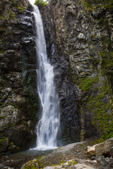 A high waterfall with white splashes of water descends from the mountain and sparkles in the sun. Attraction of Georgia Gveleti waterfall