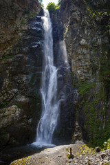 A high waterfall with white splashes of water descends from the mountain and sparkles in the sun. Attraction of Georgia Gveleti waterfall