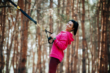 Young woman in sportswear doing exercise with TRX system in forest