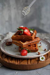 Belgium waffles with strawberry and honey at gray plate on wood board at bray background, camomile near