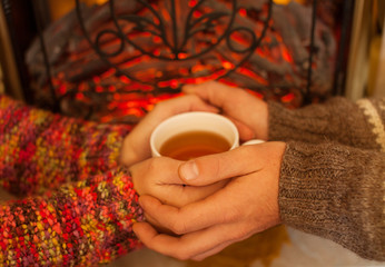 Couple hands with cup of tea near fireplace. hands of man and woman warming with help of a hot drink in mug. Couple in sweaters. 