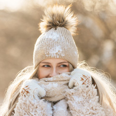 Portrait of pretty young woman with long hair in beige hat with fur pompon, warm scarf, coat, white gloves covered with snow at sunny winter day. Female wrap in and holding scarf, square cropped image