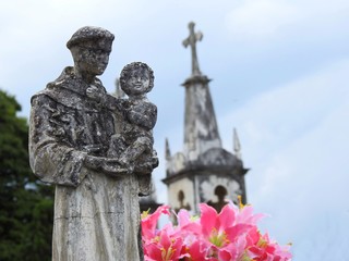 Scene in a cemetery: stone statue of a saint carrying the baby Jesus. Beside, some fake red flowers. On the blurred background, an old mausoleum.