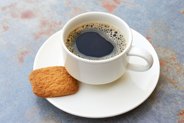 Simple white cup filled with black coffee on white saucer with brown biscuit on grey metal table with brown rust in soft light 