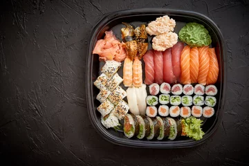Tragetasche Japanisches Essenskonzept. Catering, verschiedene Arten von Sushi auf Teller oder Plattenset. Essstäbchen, Ingwer, Sojasauce, Wasabi. Auf steinschwarzem Hintergrund in einer Kunststoffbox platziert. © Dash