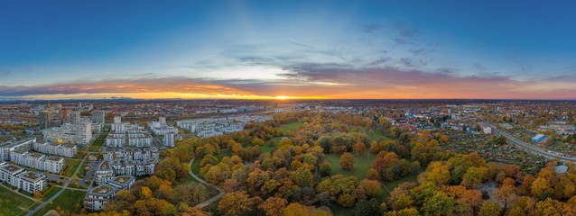 Fototapeta premium Urbanes Panorama eines Sonneruntergangs über der bayrischen Landeshauptstadt München im bunt beblätterten Hirschgarten, ein Park mit angrenzenden Wohnhäusern als Luftbild einer Drohne im Herbst