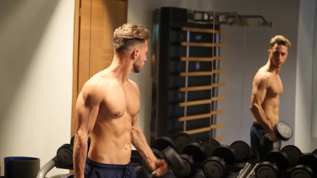 Handsome shirtless muscular young man exercising biceps in gym with dumbbells