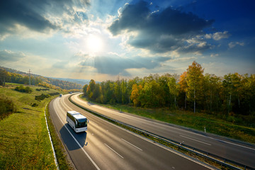 Three white buses traveling on the asphalt highway between deciduous forest in autumn colors under...