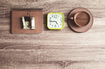 Clock, notebook, money and a cup of coffee on a wooden background. Top view