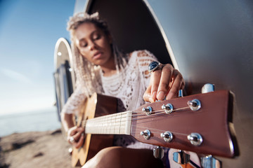 Fototapeta na wymiar Tuning the guitar. Close up of appealing inspired woman with dreadlocks tuning the guitar sitting in trailer