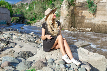 Fototapeta premium A beautiful young woman blonde with long hair in a hat sitting on a rocky shore by the river. Around the mountains and rocky terrain.