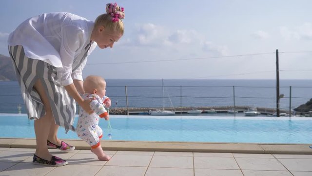 Mother supporting her little baby as he practices his first steps. The first steps of the baby near the pool on the background of the sea. 4K
