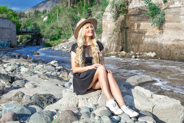 A beautiful young woman blonde with long hair in a hat sitting on a rocky shore by the river. Around the mountains and rocky terrain.