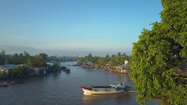 AERIAL: Flying over calm river and towards the traditional Vietnamese floating market. Idyllic view of tranquil rural life in Can Tho on a beautiful sunny evening. Large boat cruising down the river.