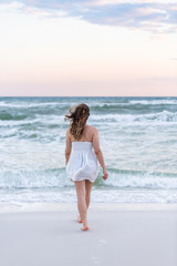 Fototapeta na wymiar Young woman in white dress on beach pink sunset in Florida panhandle with wind, ocean waves, walking legs back to water