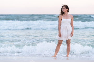 Fototapeta na wymiar Young woman front standing in white dress on beach sunset in Florida panhandle with wind, ocean waves crashing legs, tan skin, looking to side sad