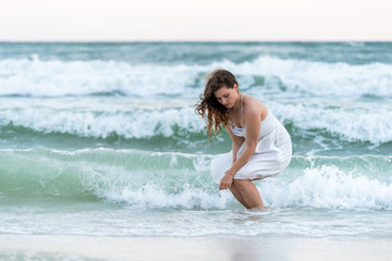 Fototapeta na wymiar Young woman sitting squatting in white dress on beach sunset in Florida panhandle with wind, ocean waves crashing legs, tan skin, looking down