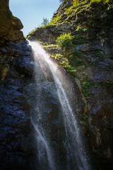 A high waterfall with white splashes of water descends from the mountain and sparkles in the sun. Attraction of Georgia Gveleti waterfall