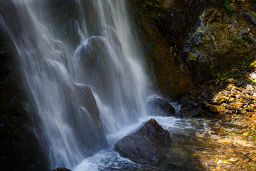 A high waterfall with white splashes of water descends from the mountain and sparkles in the sun. Attraction of Georgia Gveleti waterfall