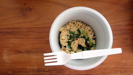 Top view of an instant cup noodle, without water and lid, on a wooden table. The dry noodle can be seen inside the cup. A plastic fork is placed on the top of the cup.