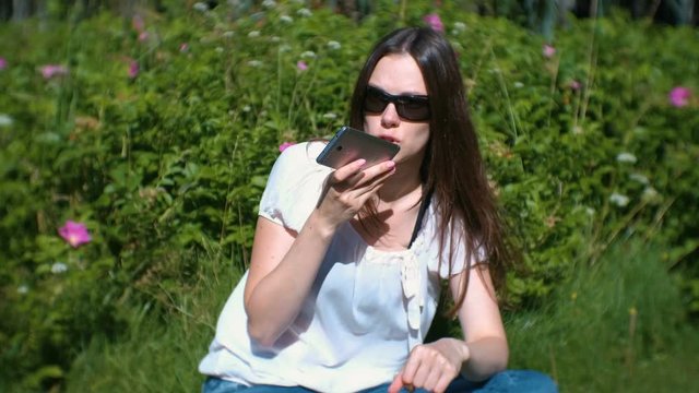 Beautiful brunette woman records a voice message on her mobile phone while sitting in the park on a Sunny day.