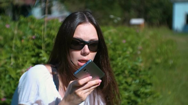 Beautiful brunette woman records a voice message on her mobile phone while sitting in the park on a Sunny day.