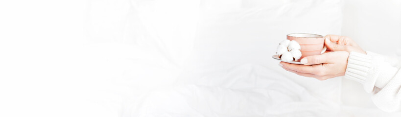 Women hands in white knitted sweater hold cup of hot tea on saucer with delicate cotton flower in the bed. Cozy morning breakfast at home. Lifestyle gentle background Copy Space autumn winter concept