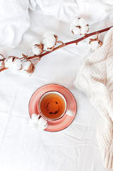 Cup of hot tea, branch of delicate cotton and knitted plaid sweater in bed with white linens. Cozy morning breakfast at home. Lifestyle gentle female background Copy Space autumn winter concept