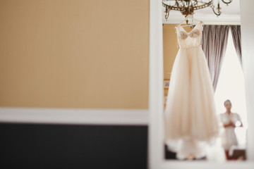 Bride looking at Stylish Wedding dress. Modern wedding gown hanging on chandelier in room. Space for text. Bride accessory for wedding. Wedding salon or showroom