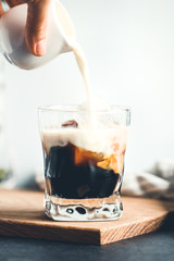 Hand is pouring milk in a glass with espresso and iced cubes.