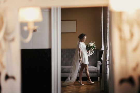 Gorgeous beautiful bride in silk robe and veil holding modern bouquet, walking to her dress. Happy stylish Bride posing near window in soft light. Mirror reflection, creative image