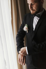 Stylish groom putting on suit and bow tie at window light. Confident and happy portrait of man. Groom getting ready in morning. Creative wedding photo