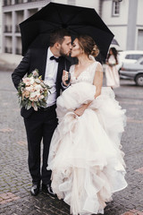 Gorgeous bride and stylish groom walking under umbrella in rainy street and kissing. Sensual...