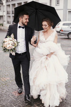 Gorgeous bride and stylish groom walking under umbrella in rainy street and smiling. Sensual wedding couple embracing. Romantic moments of newlyweds. Modern  wedding photo