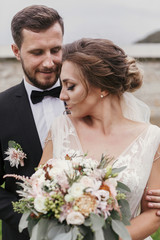 Gorgeous bride with modern bouquet and stylish groom gently hugging and smiling outdoors. Sensual...