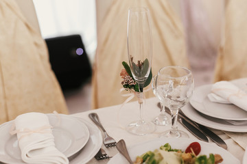 Stylish empty glasses and plate, cutlery,and floral decor on table at wedding reception in restaurant. Luxury wedding catering. Modern wedding setting
