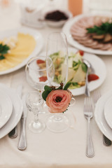Stylish empty glasses and plate, cutlery,and floral decor on table at wedding reception in restaurant. Luxury wedding catering. Modern wedding setting