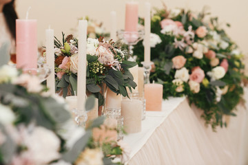 Stylish beautiful bouquets with roses and candles on pastel table at wedding reception in restaurant. Modern wedding setting. Wedding arrangements and decor