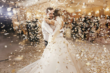 Gorgeous bride and stylish groom dancing under golden confetti at wedding reception. Happy wedding couple performing first dance in restaurant. Romantic moments - obrazy, fototapety, plakaty