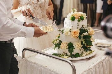 Gorgeous bride and stylish groom cutting together white wedding cake with roses at wedding...