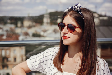 Beautiful stylish young woman in  sunglasses, retro dress and headband relaxing on roof in european city street. Hipster girl smiling and enjoying summer time in Lviv. Space for text