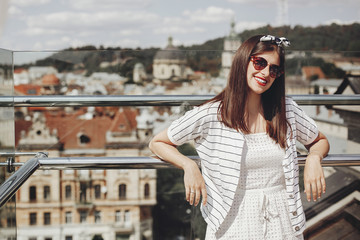 Beautiful stylish young woman in sunglasses, retro dress and headband relaxing on roof in european city street. Happy hipster girl smiling and enjoying summer time in Lviv. Copy space