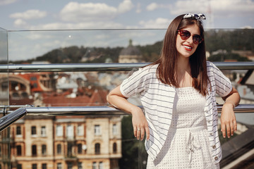 Beautiful stylish young woman in  sunglasses, retro dress and headband relaxing on roof in european city street. Hipster girl smiling and enjoying summer time in Lviv. Space for text